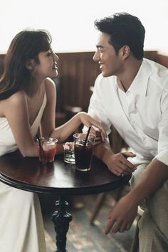 a man and woman sitting at a table with drinks in front of them, looking into each others eyes