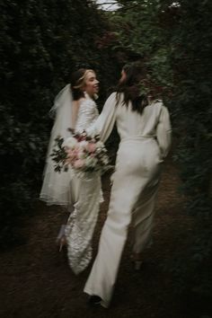 two women dressed in white are walking through the woods together and one is holding her bouquet