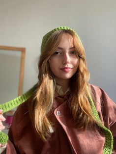 a woman with long blonde hair wearing a green knitted hat and brown coat, standing in front of a mirror