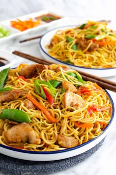 two plates with noodles, chicken and vegetables on them next to chopsticks in the background