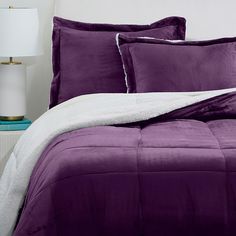 a bed with purple comforter and pillows on top of it, next to a lamp