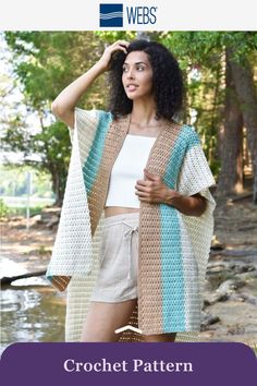 a woman wearing a crochet shawl and shorts in front of a river