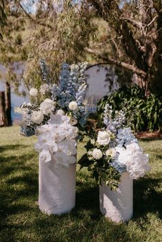 two white vases with blue and white flowers in them on the grass near some trees