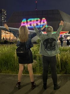 two people standing in front of a building at night with their arms up and hands raised