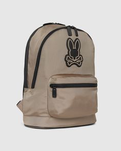 A tan MENS DOME BACKPACK - B6A628Z1BP by Psycho Bunny features a black bunny logo with crossbones on the front. It boasts a large main compartment, a padded laptop compartment, and a smaller front zippered pocket, all accented with black zippers and straps. The background is plain white. Urban Backpack, Elements Of Style, Bold Style, Bold Fashion, Weekend Getaways, Travel Style, Mens Tees, Backpack Bags, Patch Pocket