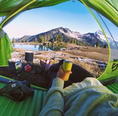 a person laying in a tent drinking from a cup with mountains in the back ground