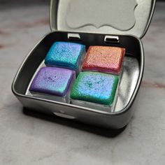 four square pieces of colored soap in a metal container on a table top with an apple logo