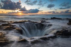 the sun is setting over an ocean with rocks and water crashing into it's shore