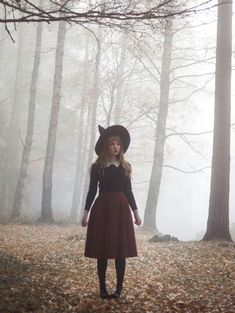 a woman standing in the woods wearing a black hat and brown dress with long sleeves