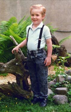 a young boy standing in front of a tree stump wearing suspenders and a white shirt