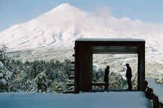 two people standing in the snow near a covered area with trees and mountains behind them