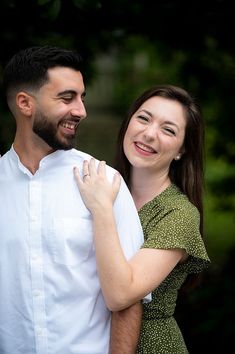 a man and woman smile as they stand close to each other in front of trees