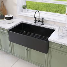 a black kitchen sink in front of a window with green cabinets and marble counter tops