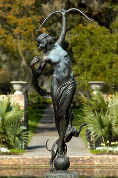 a statue of a woman holding an umbrella in front of a pond with plants and trees