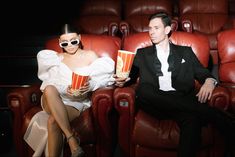 a man and woman sitting on red leather chairs holding popcorn buckets in front of them