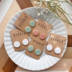 four different colored earrings on a white plate next to some flowers and a brown paper bag