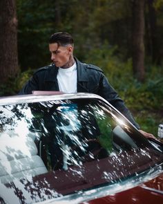 a man standing next to a red car in the woods with his hand on the windshield