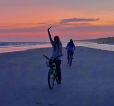 two people riding bikes on the beach at sunset with their arms up in the air