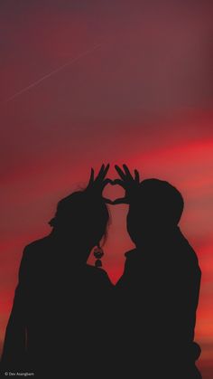 the silhouette of two people making a heart shape with their hands against an orange and pink sky