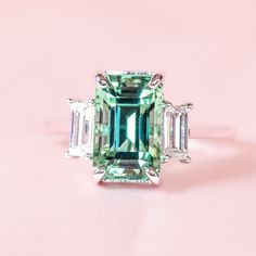 an emerald and diamond ring with three baguets on the side, sitting on a pink surface