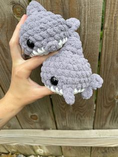 a hand holding two gray crocheted stuffed animals in front of a wooden fence