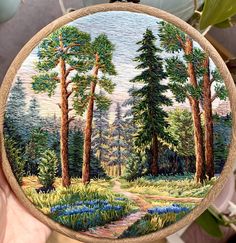 a person holding up a small embroidery art piece with trees and flowers in the background