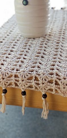 a crocheted table runner with tassels on it and a white vase in the background