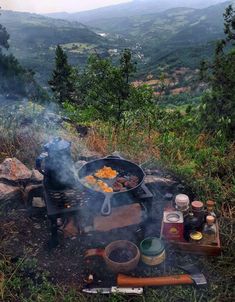 an outdoor grill with food cooking on it in the mountains near some trees and grass