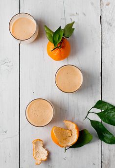 three glasses filled with orange juice on top of a white wooden table next to leaves