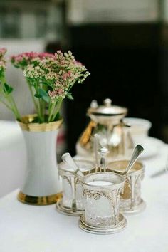 three silver cups with spoons are sitting on a table next to a vase filled with flowers