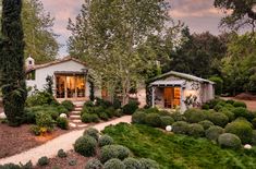 a white house surrounded by lush green trees and shrubbery in front of it is lit up at night