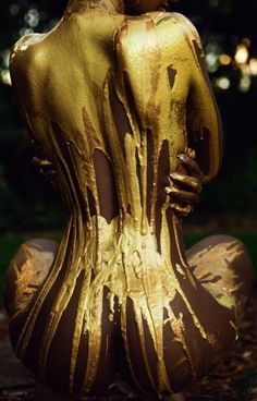 a woman covered in gold paint sitting on the ground with her back to the camera