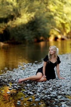 a woman sitting on the ground next to a body of water with rocks in front of her