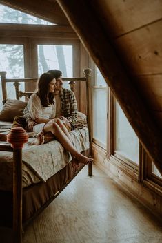 a man and woman sitting on a bed in a room with wooden walls, windows, and wood flooring