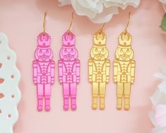 three pairs of pink and gold robot earrings on a pink background with white flowers in the foreground