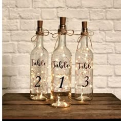 three bottles with lights in them sitting on top of a wooden table next to a brick wall