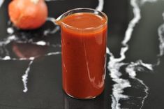 a glass filled with red liquid next to an orange on a black counter top and another orange in the background