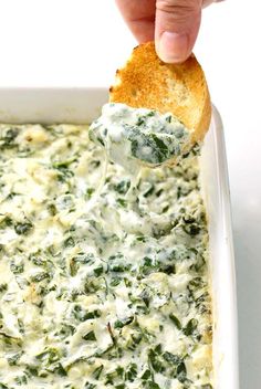 a hand dipping a piece of bread into a white dish filled with spinach dip