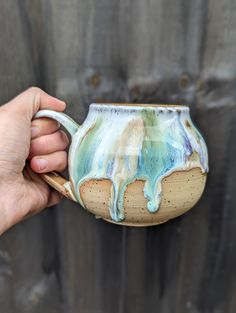 a hand holding a ceramic mug with blue and green glaze on the outside, in front of a wooden fence