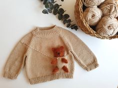 a knitted sweater with a teddy bear on it next to some balls of yarn