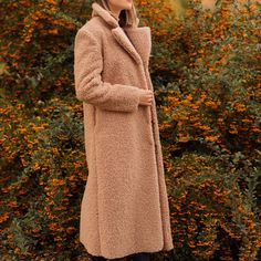 a woman standing in front of bushes wearing a tan coat and black boots with her hands on her hips