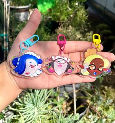 a hand holding three different colored cartoon keychains in front of some succulent plants