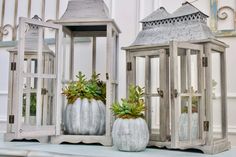 three white lanterns with plants in them sitting on a table