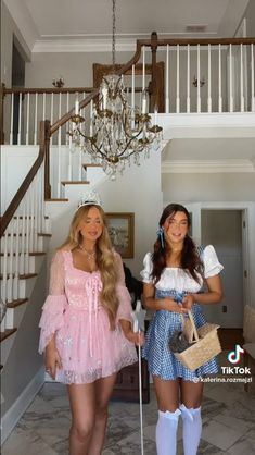 two women dressed in costume standing next to each other near stairs and chandelier