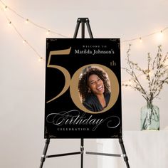 a black and gold 50th birthday party sign with a photo on it, in front of a string of lights