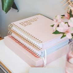 a stack of pink notebooks sitting on top of a table next to a vase with flowers