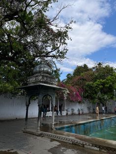 a gazebo in the middle of a pool surrounded by trees and people walking around