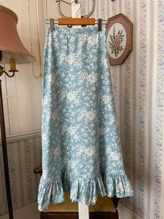 This long skirt is made from a soft cotton blend in light sky blue with an overall white floral pattern. It has a ruffle at the bottom and fastens at the back with two hook and eye closures and a short zipper. There are no labels. The measurements, taken with the skirt lying flat, are: waist, 12 inches; hips, 19 inches; length, 33 1/2 inches; bottom edge, 38 inches. In very good vintage condition. Spring Maxi Skirt With Ruffle Hem, Light Blue Cotton Tiered Skirt, Light Blue Tiered Cotton Skirt, Spring Cotton Maxi Skirt, Blue Ruffled Maxi Skirt, Light Blue Flowy Cotton Skirt, Light Blue Tiered Maxi Skirt For Summer, Fitted Light Blue Maxi Skirt For Spring, Spring Blue Ruffled Maxi Skirt