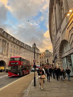 people are walking on the sidewalk in front of a red double decker bus and buildings