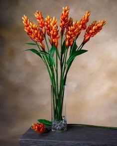 an arrangement of orange flowers in a glass vase on a wooden table against a beige background